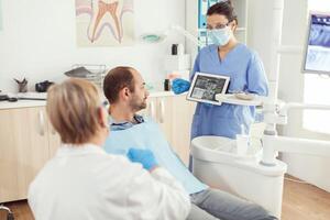 Stomatology nurse in dentistiry office showing tablet pc with tooth radiography explaining toothache treatment. Man sick patient sitting on dental chaiar in hospital orthodontist clinic office photo