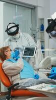 Dentist in protective equipment showing on tablet dental x-ray reviewing it with senior patient. Medical team wearing face shield coverall, mask, gloves, explaining radiography using notebook display photo