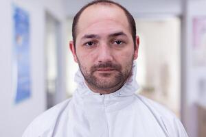 Portrait of exhausted doctor in office looking on camera wearing ppe suit without face shield sitting on chair in waiting room clinic. Concept of new normal medic in coronavirus outbreak photo