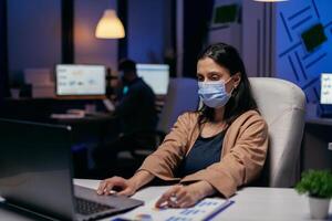 empleado con proteccion cara máscara trabajando tarde a noche en nuevo normal oficina. mujer siguiendo social distanciamiento reglas debido a coroanvirus pandemia mientras trabajando tarde horas a el oficina. foto