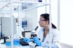 Laboratory technician working in microbiology lab using computer and microscope. Chemist wearing lab coat using modern technology during scientific experiment in sterile environment. photo