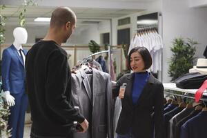 Fashion boutique arab customer asking asian woman assistant for opinion while choosing formal jacket. Clothing store seller and buyer holding apparel on hanger and discussing style photo