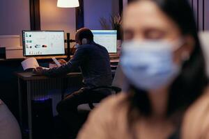 Man wearing face mask working on deadline as safety precaution against covid19 outbreak. Woman following social distancing rules due to coroanvirus pandemic while working late hours at the office. photo