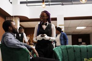 Friendly African American female waitress taking order serving hotel guest in lobby. Young black man traveler ordering coffee while waiting for check-in. Customer service in hospitality industry photo