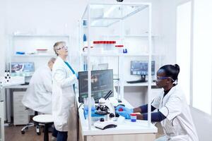 científico con africano etnia participación muestra recipiente trabajando con su equipo en moderno instalación. negro cuidado de la salud investigador en bioquímica laboratorio vistiendo estéril equipo. foto