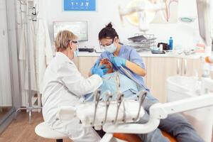 Sick patient sitting on stomatology chair while medical nurse looking into mouth analyzing teeth health during stomatology examination. Senior dentist woman waiting for stomatological procedure photo