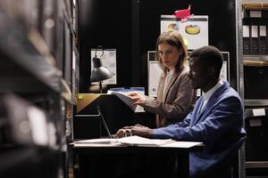 Depository workers reading administrative files, discussing bureaucracy record in storage room. Diverse bookkeepers working overtime at accountancy report, analyzing management research photo