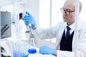 Experienced scientist working in biotechnological laboratory with sterile dropper and blue liquid. Senior professional chemist using pippete with blue solution for microbiology tests. photo