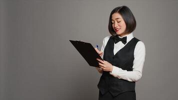 Asian hotel concierge creating a checklist of registrations, working on online reservations and bookings in studio. Receptionist looking at clipboard papers over grey background. Camera A. photo