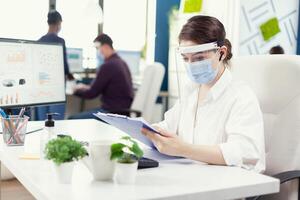 Financial advisor with wireless headphones checking financial statistic in busy office with face mask. Employees with visors working in corporate company workspace respecting social distance analysing data and graphs. photo