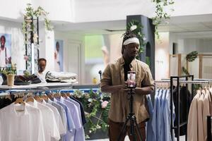 africano americano influenciador promover ropa Tienda mientras grabación vídeo en teléfono inteligente hombre creando en línea social medios de comunicación publicidad en móvil teléfono en compras centro comercial Moda boutique foto