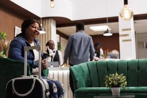 Happy young African American woman sitting at hotel lobby with suitcase holding smartphone and smiling, using phone for mobile self check-in. Black female tourist with luggage waiting for room photo