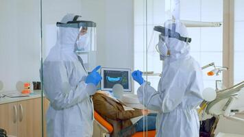 Team of doctors wearing full virus protection uniform standing near patient analysing teeth digital x-ray using tablet planning treatment. Concept of new normal dentist visit in coronavirus outbreak. photo