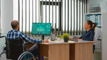 Paralysed financial worker sitting in wheelchair immobilized analysing economy statistics of company in business office discussing with colleague. Handicapped businessman using modern technology photo