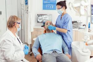 Sick man preparing for tooth surgery while nurse putting medical oxigen mask sitting on dental chair during stomatology hospital office. Dentist senior woman examining toothache treatment photo