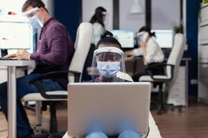 African freelancer in workplace using laptop wearing face mask against covid19. Multiethnic business team working respecting social distance during global pandemic with coronavirus. photo