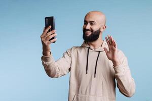 sonriente árabe hombre ondulación Hola mientras hablando en línea utilizando teléfono inteligente videollamada solicitud. simpático persona teniendo remoto comunicación, saludo con mano y Hablando con móvil teléfono frente cámara foto