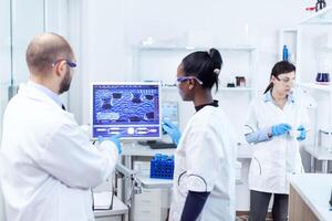 African scientist and coworker discussing their research about virus in busy lab. Multiethnic team of medical researchers working together in sterile lab wearing protection glasses and gloves. photo