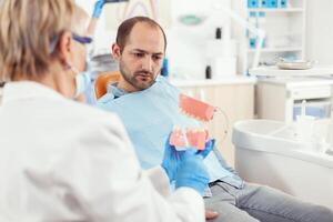 Stomatologist explaining proper dental hygiene using teeth skeleton during stomatology appointment. Dentist preparing sick man patient for dentistry surgery sitting on chair in examination room photo