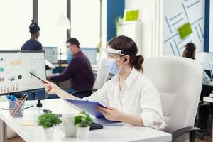 Businesswoman looking at financial stats wearing face mask during covid-19. Employees with visors working in corporate company workspace respecting social distance analysing data and graphs. photo