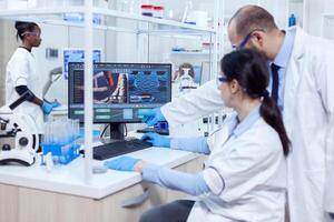 Group of scientist working with computer in laboratory. Team of researchers doing pharmacology engineering in sterile lab for healthcare industry with african assistant in the background. photo