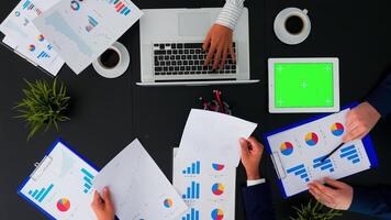 Top view of unrecognisable businessman looking on green screen of tablet during brainstorming in conferance room analysing graphs with coworkers sitting on office table. Copy space, flat lay concept photo