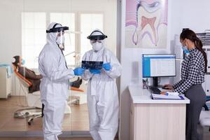 Stomatology team dressed up in ppe suit during global pandemic with coronavirus in dental reception holding patient x-ray, keeping social distancing. Receptionist with face mask as prevention. photo