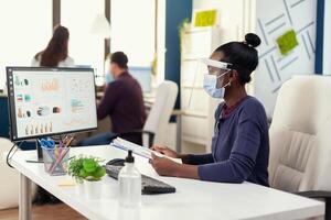 African businesswoman checking financial analysis on computer holding clipboard wearin face mask during coronavirus. Multiethnic coworkers working respecting social distance in financial company. photo