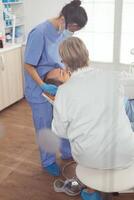 Sick patient sitting on stomatological chair with open mouth during dental examination. Senior woman stomatologist and medical nurse with masks holding sterilized tools checking teeth health photo