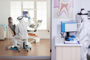 Stomatologist in protective suit asking for patient dental x-ray examining teeth problems during coronavirus pandemic in modern clinic. Medical team wearing coverall, face shield, mask and gloves. photo