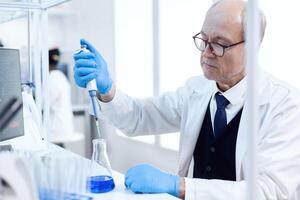 Elderly scientist doing genetic experiment in busy laboratory using sterile dropper with blue liquid. Senior professional chemist using pippete with blue solution for microbiology tests. photo