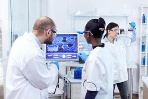 africano científico y médico estudiando cura para peligroso virus en computadora en ocupado laboratorio. multiétnico equipo de médico investigadores trabajando juntos en estéril laboratorio vistiendo proteccion lentes y guantes. foto