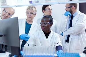 Team of scientists with multiethnicity discussing chemical formula in front of computer. Black healthcare researcher in biochemistry laboratory wearing sterile equipment. photo