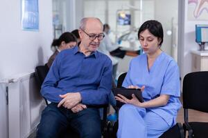 Nurse taking notes on tablet pc discussing with senior patient in dental office, explaining diagnosis. Assistant filling writing healthcare form. Dentistiry hospital. photo