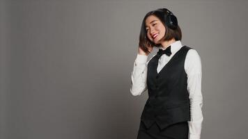 Hotel personnel listening to music using audio headset, having fun with groovy mp3 tunes over grey background. Asian receptionist enjoying songs on radio with headphones in studio. Camera A. photo