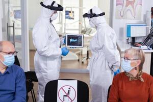 Man discussing with nurse in dental reception wearing protection suit against coronavirus, elderly patients waiting in reception keeping distance. Concept of new normal dentist visit in outbreak. photo