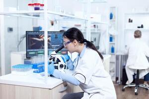 Expert in genetics doing research using microscope to analise chemical from human adn. Chemist wearing lab coat using modern technology during scientific experiment in sterile environment. photo