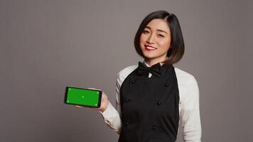 Catering employee holding phone with greenscreen display, pointing at chromakey design on smartphone. Asian waitress using isolated copyspace template over grey background in studio. Camera B. photo