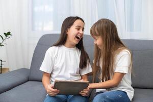 Young twins using a laptop and a tablet sitting on a couch in the living room photo