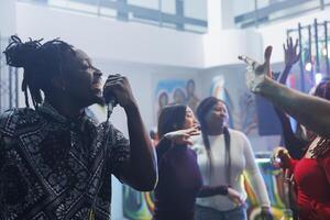 Smiling cheerful african american man singing in microphone while clubbing with friends. Young carefree clubber partying, holding mic and enjoying karaoke in crowded nightclub photo