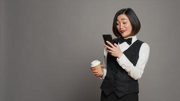 conserje personal Bebiendo taza de café en estudio, hojeada teléfono inteligente social medios de comunicación aplicaciones asiático recepcionista disfrutando cafeína refresco mientras ella usos móvil teléfono. cámara una. foto