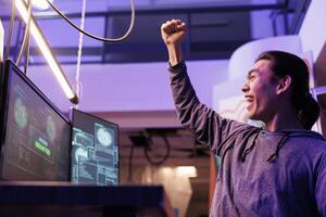 Happy hacker making victory gesture after getting server access granted. Cheerful excited asian man celebrating hacking computer network system successfully and raising fist photo