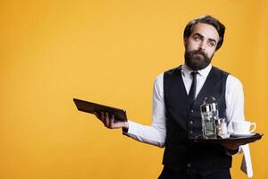 Skilled employee gives menu on tablet to customers at five star restaurant, waiter prepares meal prices while he serves clients. Butler with suit and tie working as professional catering personnel. photo