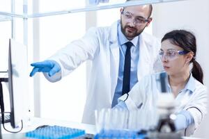 Young female scientist working with her male supervisor usinc omputer in busy laboratory. Team of researchers doing pharmacology engineering in sterile lab for healthcare industry. photo