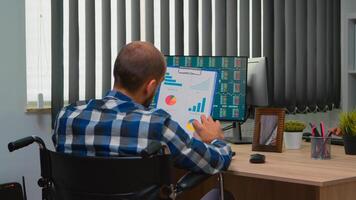 Handicapped businessman working with financial statistics checking graphs sitting immobilized in wheelchair on desk in building office. Paralysed freelacer using modern technology and documents. photo