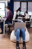 African employee with protection mask against covid-19 pandemic sitting on armchair in middle of office room holding digital tablet. Multiethnic business team working respecting social distance during global pandemic with coronavirus. photo