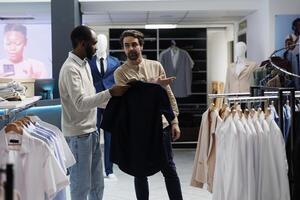 africano americano hombre participación camisa en percha y preguntando compras centrar trabajador acerca de Talla opciones boutique trabajador señalando a nuevo colección vestir estante, Ayudar cliente a escoger atuendo foto