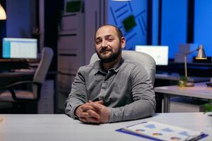 Portrait of concentrated manager smiling at camera thinking about success. Smart businessman sitting at his workplace in the course of late night hours doing his job. photo