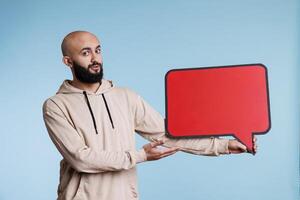 Arab man presenting product with empty speech bubble for promotional message mock up. Young arabian person holding blank dialog frame with copy space and looking at camera photo