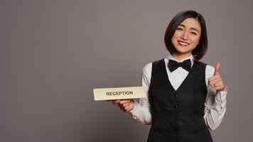 Hotel concierge holding sign to indicate direction for reception desk, presenting indicator to help guests and offer assistance in studio. Employee with uniform and bow uses wall pointer. Camera B. photo